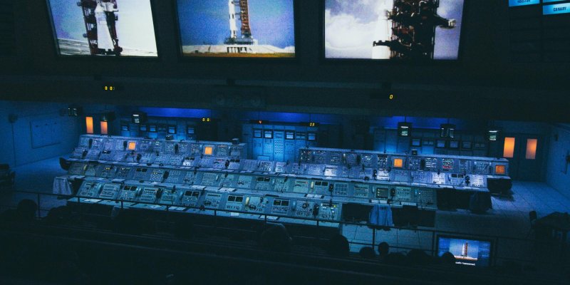 Kontrollraum für einen Raketenstart. Im Hintergrund Bildschirme mit Bildern der Startrampe und Rakete. Im Vordergrund viele Kontrollstationen und Messinstrumente.